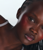A close-up of a woman with dark skin and braided hair, wearing a gray strap top. She has glossy lips, bold red blush, and acne-prone skin using the Hydrocolloid Breakout Recovery Mask, gazing thoughtfully against a light background.